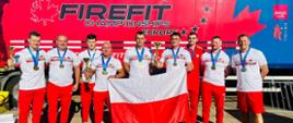 Dziewięciu zawodników z Polski pozuje z medalami, pucharami i biało-czerwoną flagą przed czerwono-niebieską ciężarówką z dużym logiem FireFit Championships Europe i stylizowanym liściem klonu.