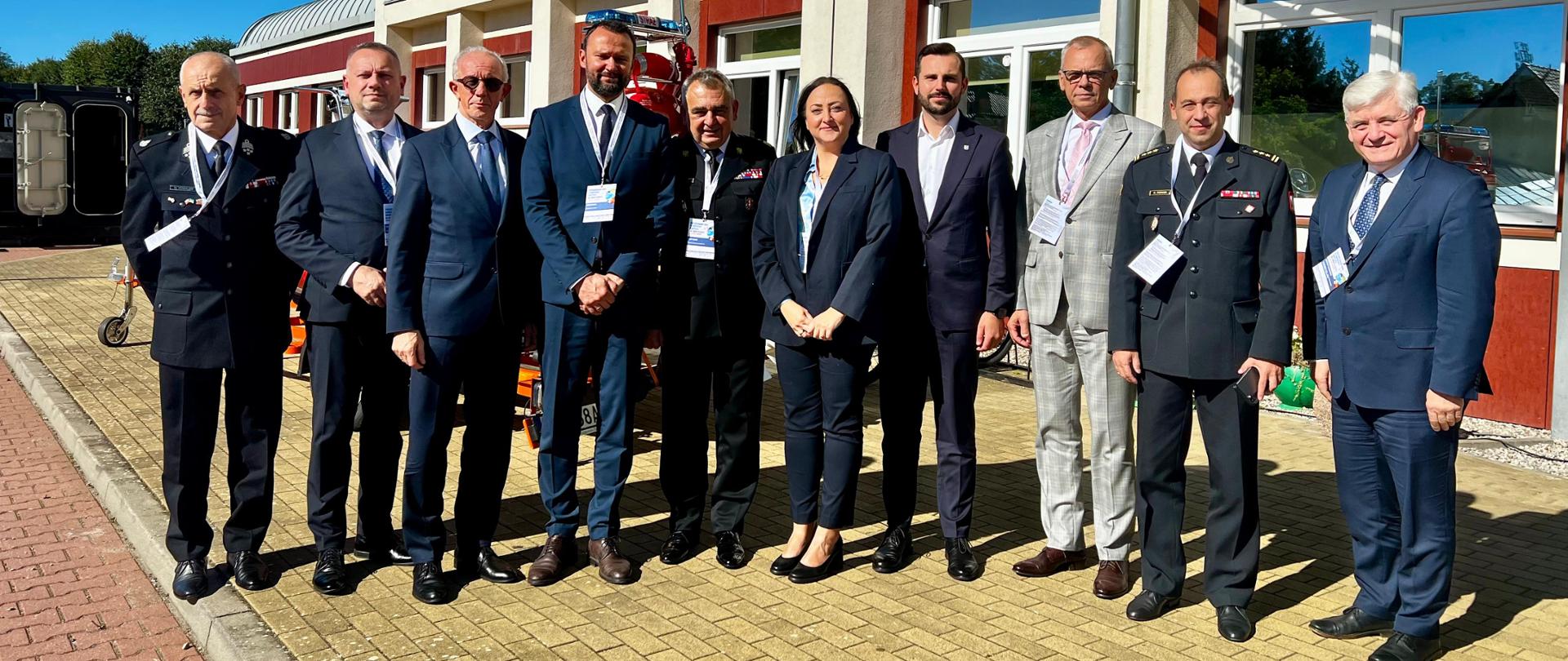 Zdjęcie grupowe uczestników konferencji przed budynkiem. W pierwszym rzędzie stoją przedstawiciele Państwowej Straży Pożarnej w mundurach galowych oraz osoby w garniturach i garsonce. Wszyscy pozują do wspólnej fotografii w pełnym słońcu.