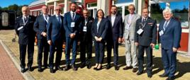 Zdjęcie grupowe uczestników konferencji przed budynkiem. W pierwszym rzędzie stoją przedstawiciele Państwowej Straży Pożarnej w mundurach galowych oraz osoby w garniturach i garsonce. Wszyscy pozują do wspólnej fotografii w pełnym słońcu.
