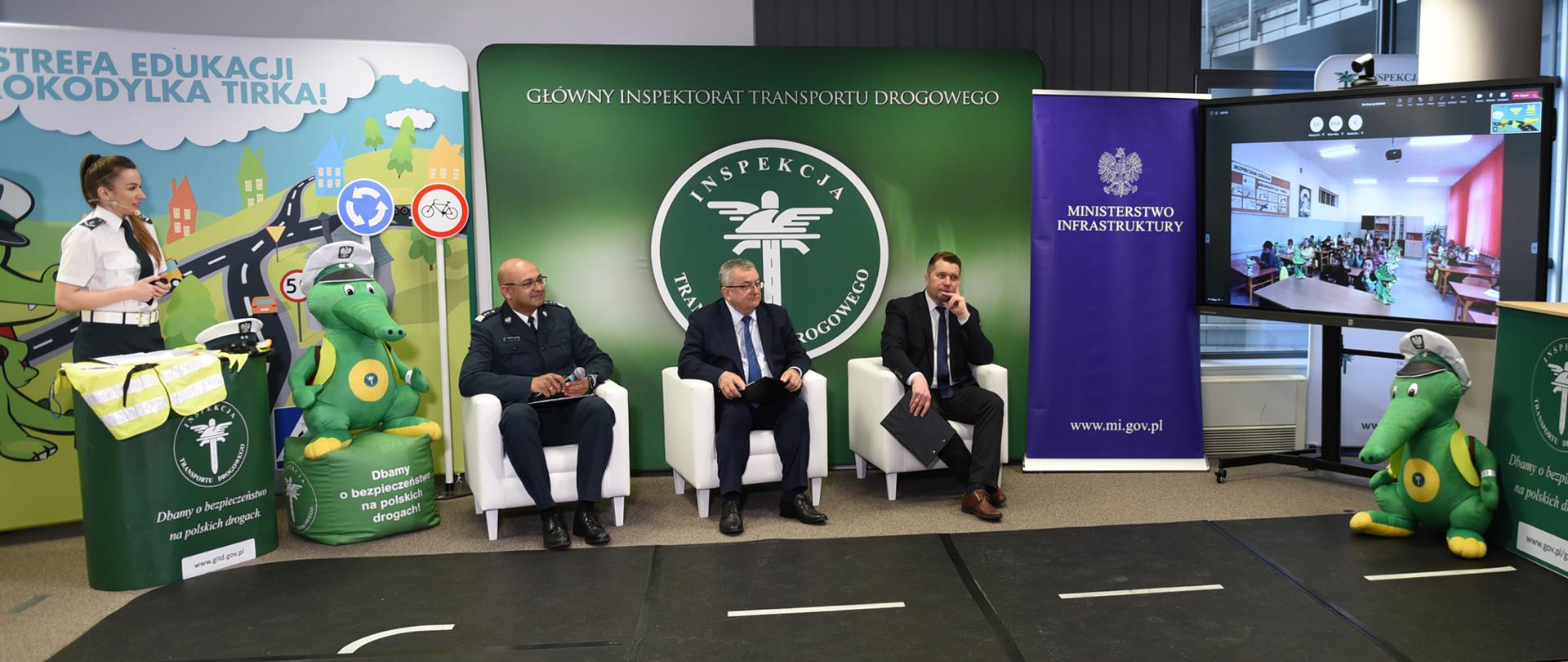 Lekcja bezpieczeństwa ruchu drogowego online dla uczniów z Ukrainy. Od lewej siedzą: Główny Inspektor Transportu Drogowego Alvin Gajadhur, Minister Infrastruktury Andrzej Adamczyk i Minister Edukacji i Nauki Przemysław Czarnek. Po prawej stronie monitor i wyświetlony na nim obraz z sali lekcyjnej.