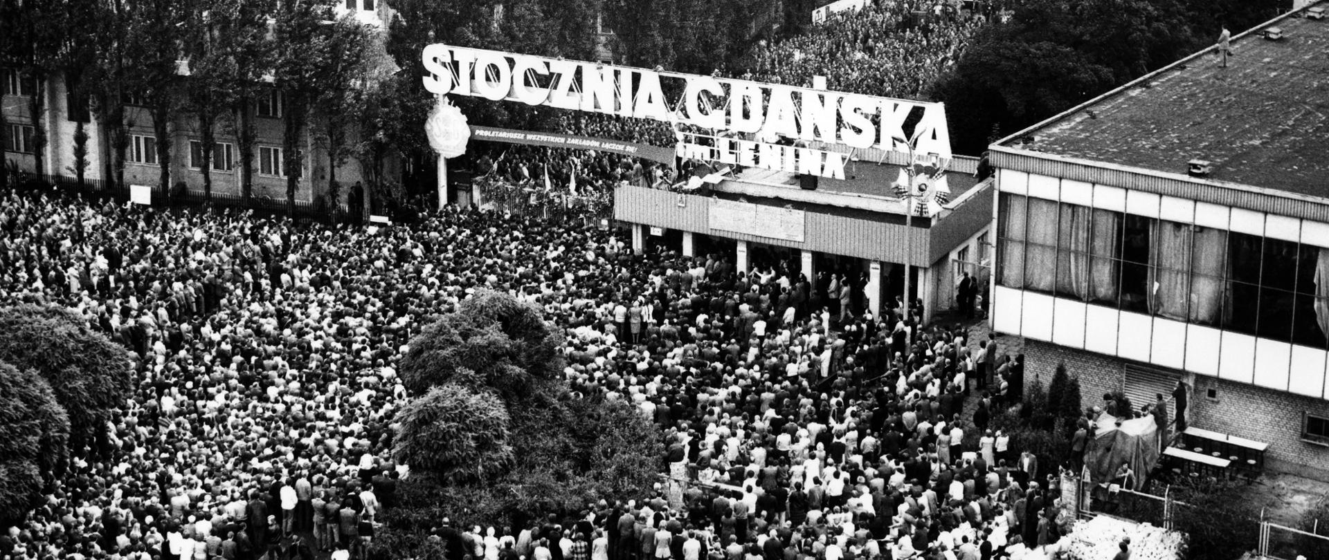 Серпнева страйк на Ґданській верфі ім. Леніна. Вид зверху на Ворота № 2 та меканців, які зібралися під нею та працівники верфі. Над воротами розміщено транспарент з гаслом "ПРОЛЕТАРИ ВСІХ ЗАВОДІВ, ЄДНАЙТЕСЯ!", а на даху караулу - дошки з 21 постулатами.