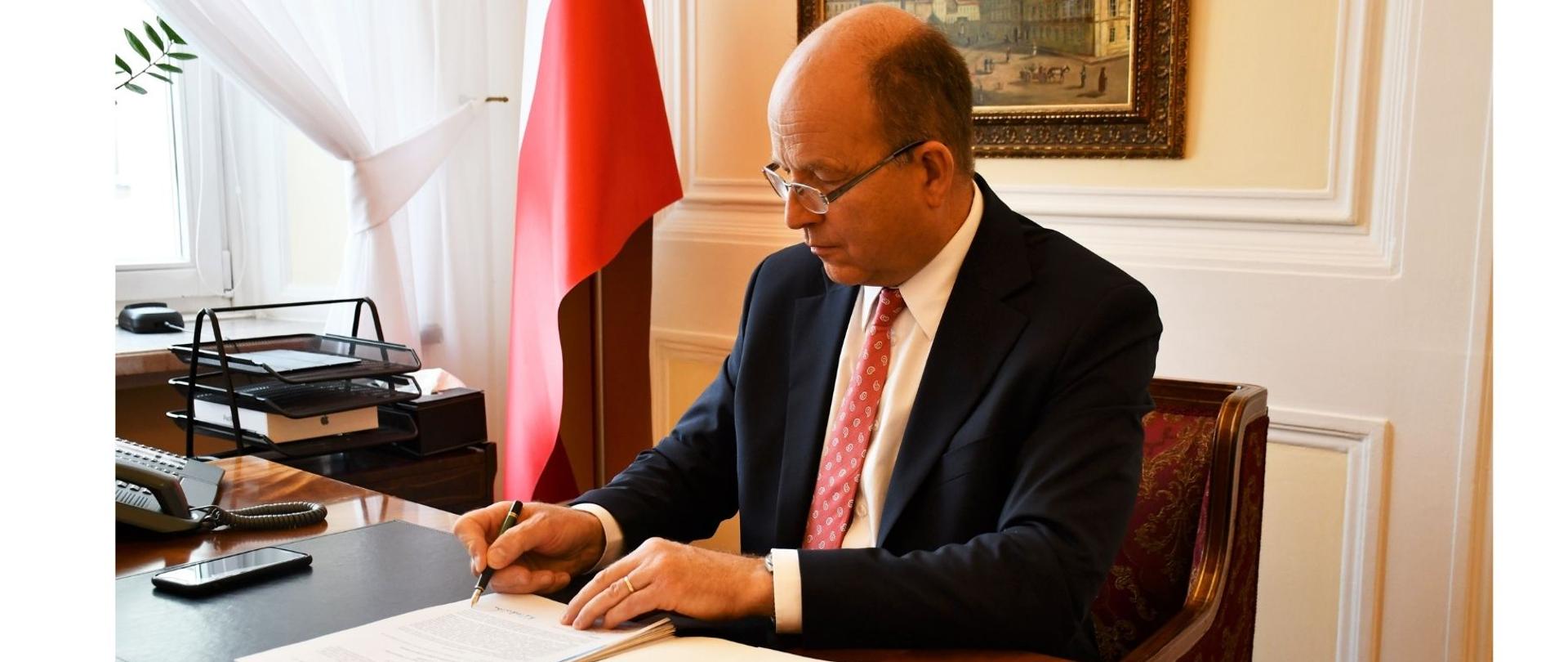 Na zdjęciu znajduje się elegancko ubrany mężczyzna. Mężczyzna ten siedzi za biurkiem, przy którym podpisuje dokument. W tle znajduje się biało-czerwona flaga, obraz. 