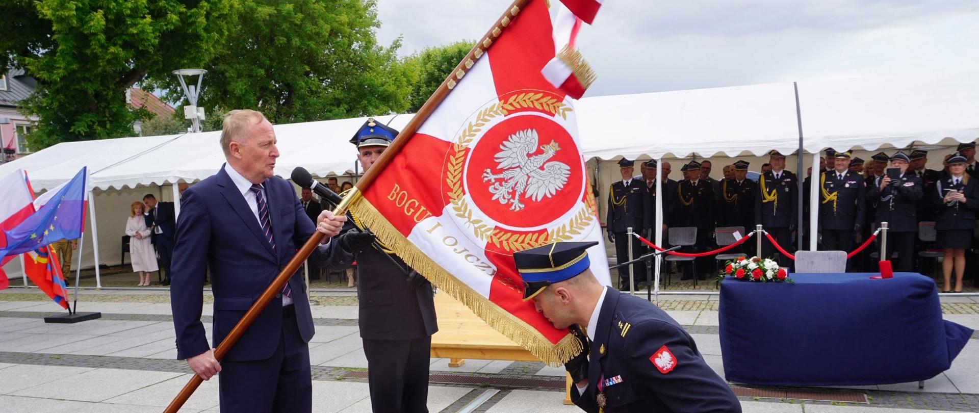Sekretarz Stanu w MSWiA trzyma sztandar Komendy Powiatowej PSP w Węgrowie, a klęczący funkcjonariusz Państwowej Straży Pożarnej oddaje mu cześć. Uroczystość odbywa się na czerwonym dywanie przed trybuną honorową.
