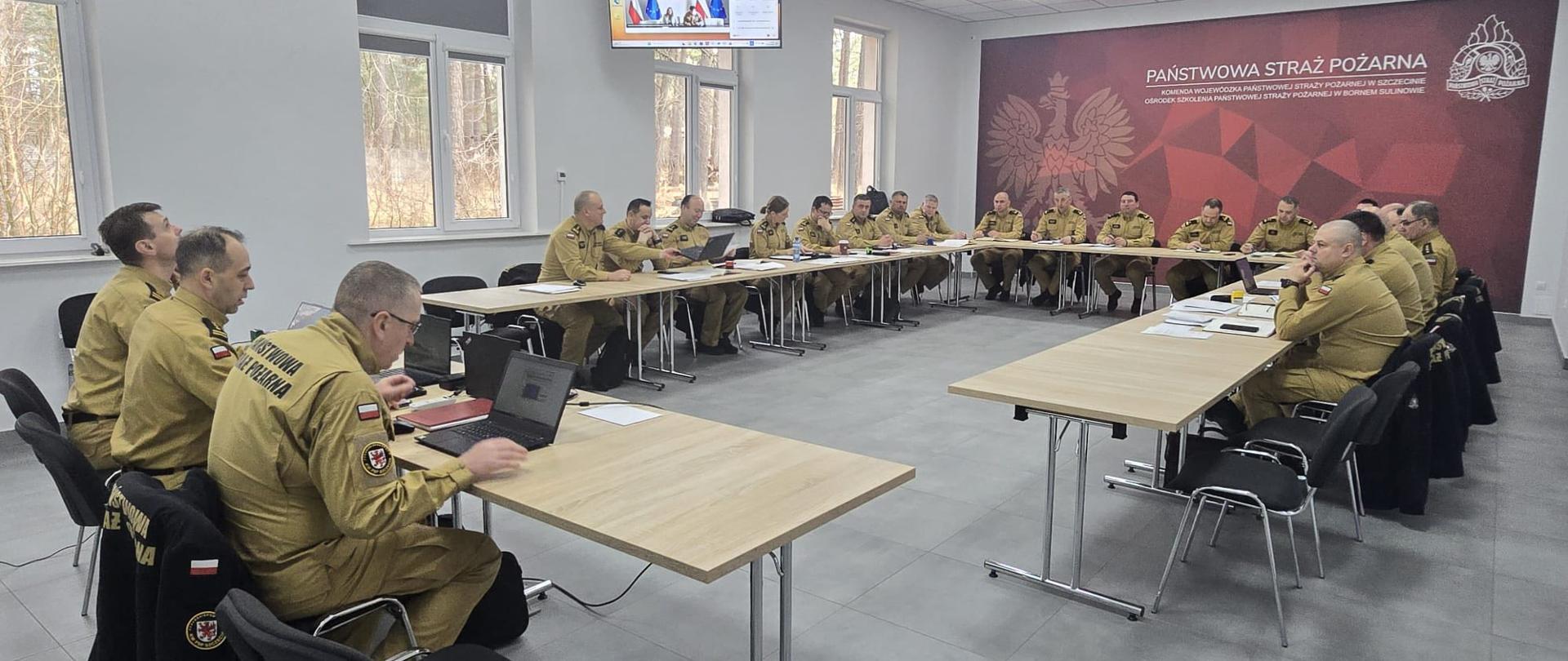 Zdjęcie przedstawia jasną, nowoczesną salę konferencyjną. W pomieszczeniu ustawiono stoły w kształcie litery „U”. Przy stołach siedzi kilkanaście osób w jednakowych, jasnobrązowych mundurach strażackich. Większość z nich to mężczyźni. Uczestnicy spotkania siedzą na krzesłach, część korzysta z laptopów lub notatek, inni słuchają.
Na tylnej ścianie znajduje się duża, czerwona plansza z napisem „Państwowa Straż Pożarna” oraz symbolem orła. Nad stołem, na środku sali, zawieszony jest ekran, na którym wyświetlana jest prezentacja lub wideokonferencja.
Sala jest dobrze oświetlona światłem sufitowym oraz dziennym wpadającym przez duże okna po lewej stronie. Za oknami widać drzewa. Podłoga jest jasnoszara, a ściany białe.