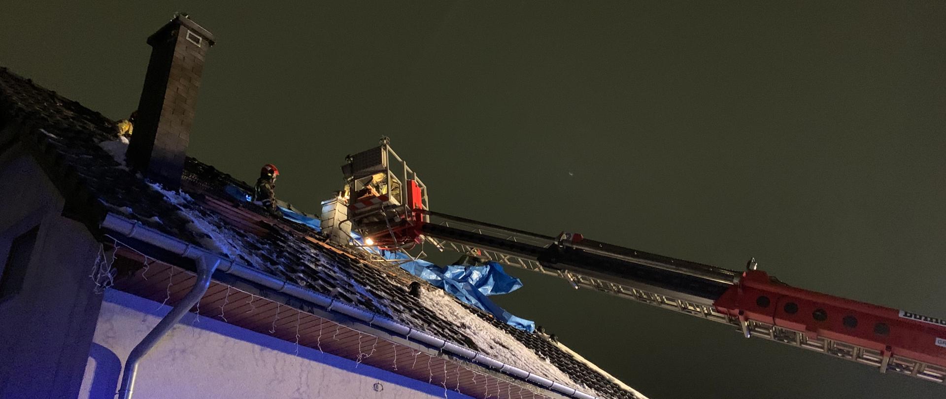 Zdjęcie zrobione w nocy przedstawia działania strażaków po pożarze poddasza budynku jednorodzinnego. Zdjęcie przedstawia budynek jednorodzinny z częściowo spalonym dachem. na poddaszu ponad konstrukcją dachu widać trzech strażaków PSP w piaskowych ubraniach i czerwonych hełmach zabezpieczających plandeką zniszczony dach. Nad dachem widać wysięgnik pożarniczego podnośnika. W koszu podnośnika znajduje się czwarty strażak pomagający w pracach zabezpieczających. 