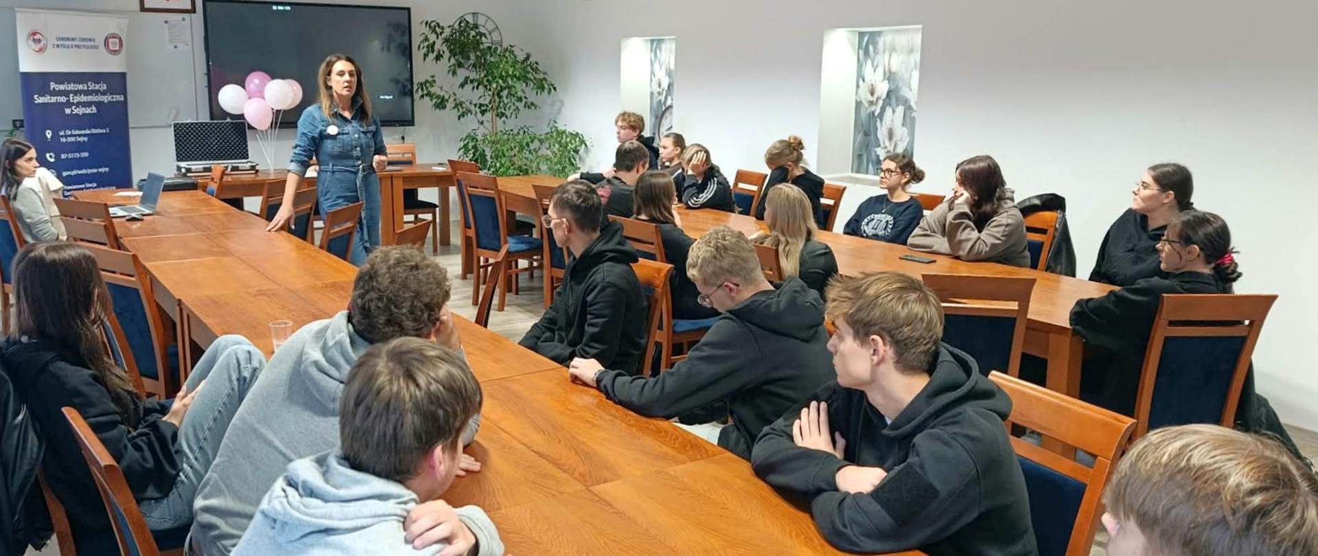 Młodzież szkolna słucha prelekcji prowadzonej przez kobietę.