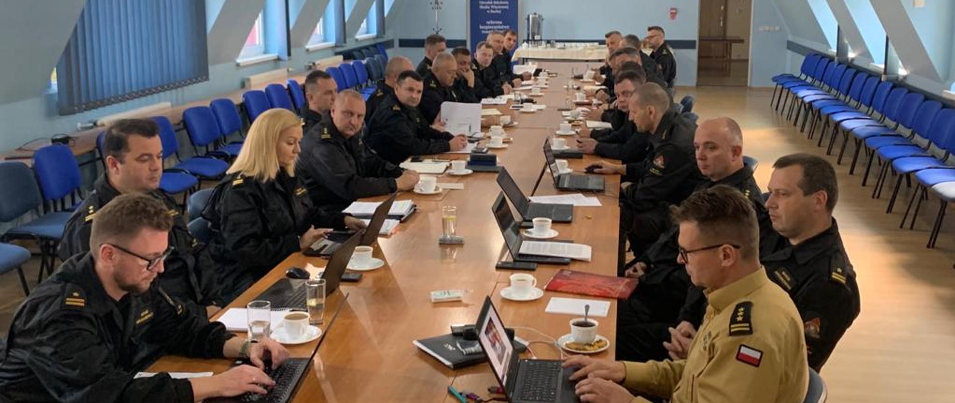 Przy długim stole , w sali konferencyjne, z obu stron siedzą funkcjonariusze PSP. Na stole materiały piśmienne, sprzęt komputerowy. Uczestnicy w liczbie ok. 30 osób. Umundurowaniu w mundury dowódczo sztabowe.