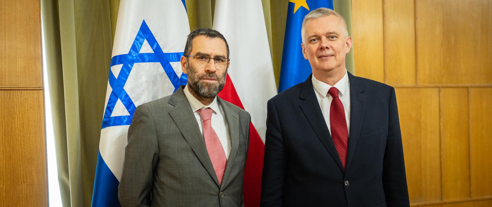 Dwóch mężczyzn w formalnych garniturach stoi obok siebie i patrzy w obiektyw aparatu. Mężczyzna po lewej ma ciemne włosy z siwizną, brodę, okulary oraz szary garnitur z białą koszulą i czerwonym krawatem. Mężczyzna po prawej ma krótkie siwe włosy i ubrany jest w granatowy garnitur, białą koszulę oraz czerwony krawat. W tle znajdują się flagi Izraela, Polski i Unii Europejskiej oraz jasne zasłony i drewniane panele.