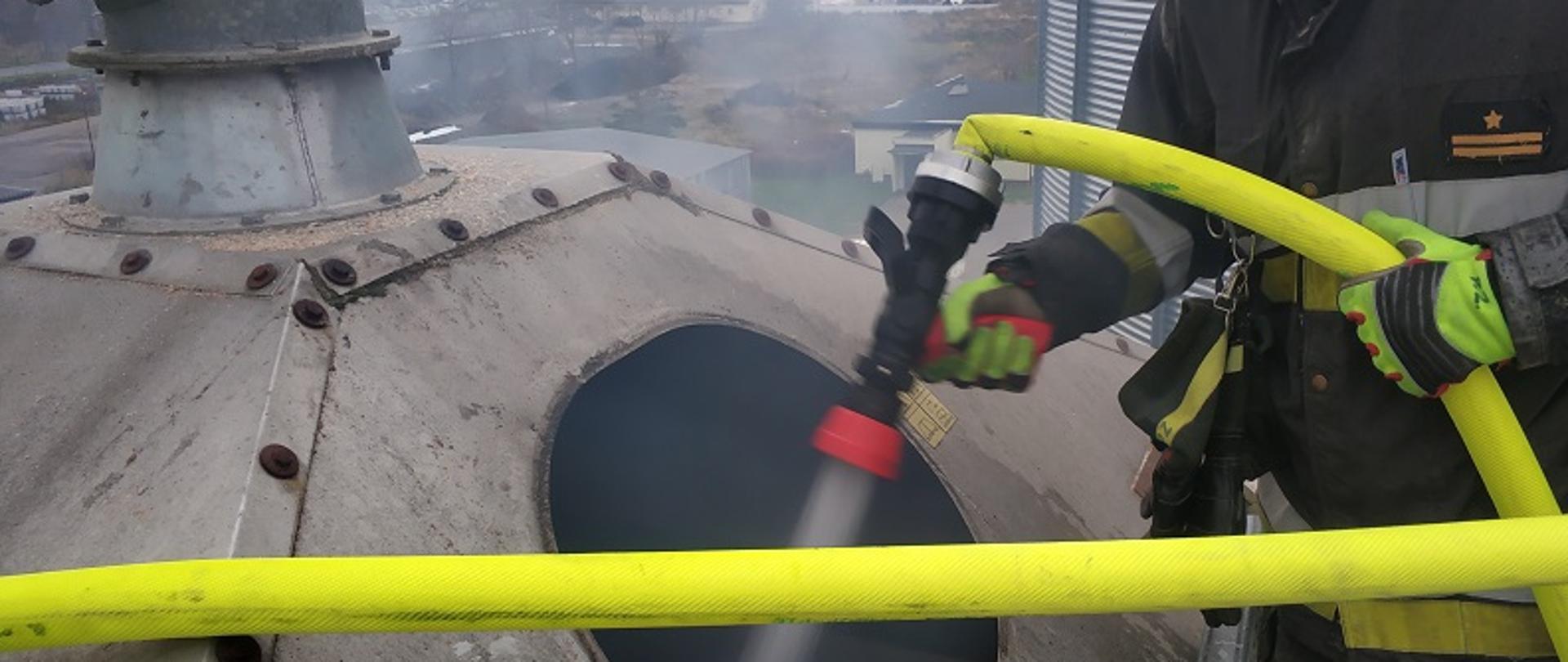 Zdjęcie przedstawia strażaka w ubraniu specjalnym który za pomocą węża i prądownicy wodnej gasi wnętrze komory suszarnianej przez otwór technologiczny