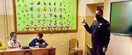 Zdjęcie przedstawia strażaka podczas prowadzenia zajęć edukacyjnych. W tle widać tablice edukacyjne, strażak w maseczce i umundurowany. W ławkach widać dzieci.