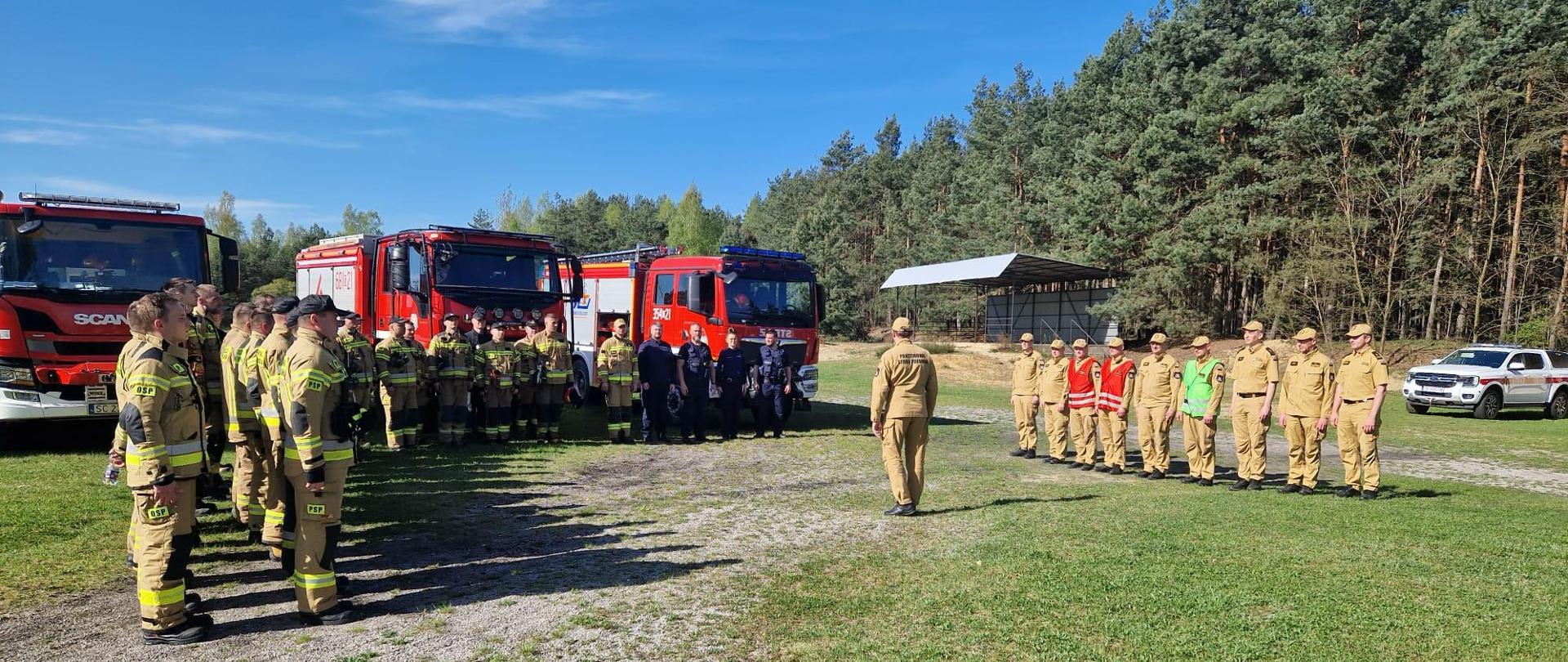 Na zdjęciu strażacy i policjanci podczas odprawy na placu ćwiczeń stojący w dwuszeregu w centralnym punkcie dowódca ćwiczeń wydający rozkazy dla poszczególnych grup, w tle samochody strażackie oraz las.