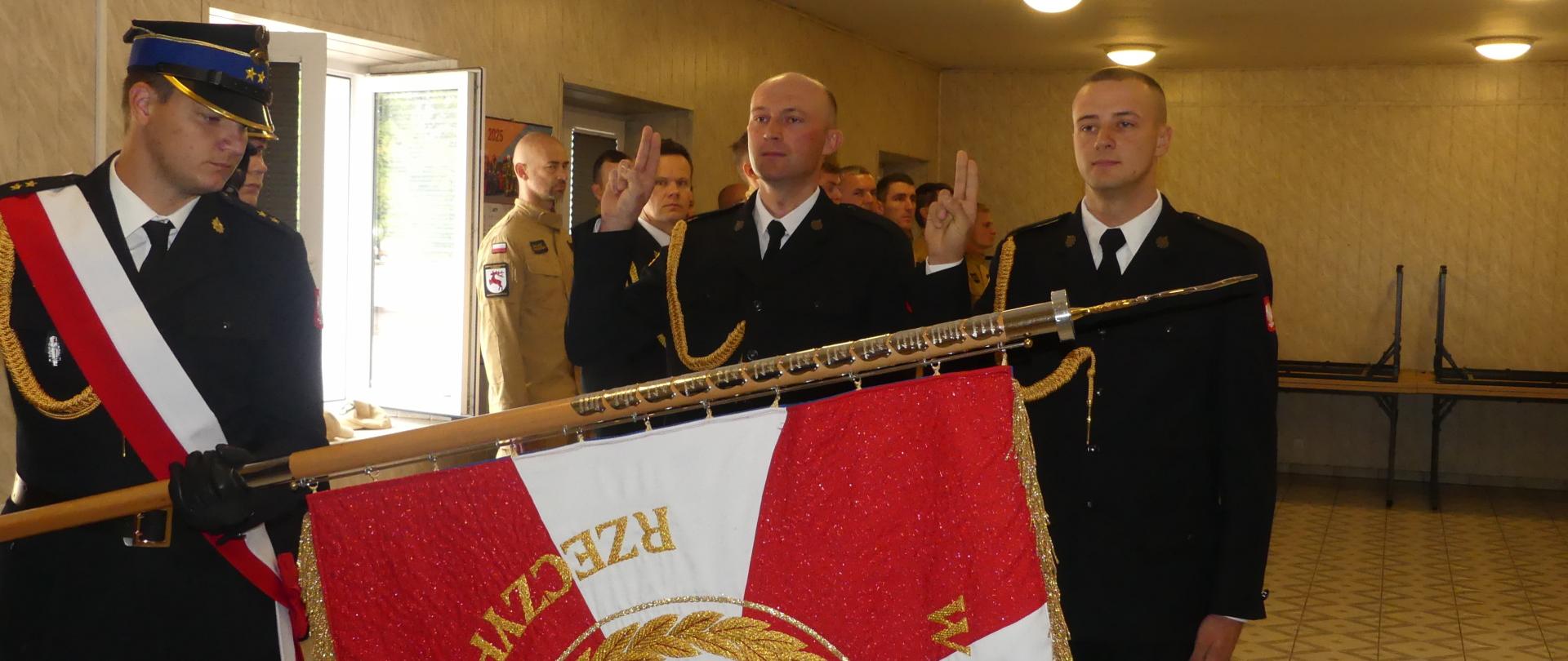 Zdjęcie przedstawia dwóch nowych strażaków Komendy Powiatowej podczas uroczystości ślubowania - str. Jakub Sikora oraz str. Marcina Sobierajskiego. Ślubujący składają przysięgę unosząc dwa palce prawej dłoni. Przed nimi strażak w mundurze galowym pochyla sztandar Komendy.