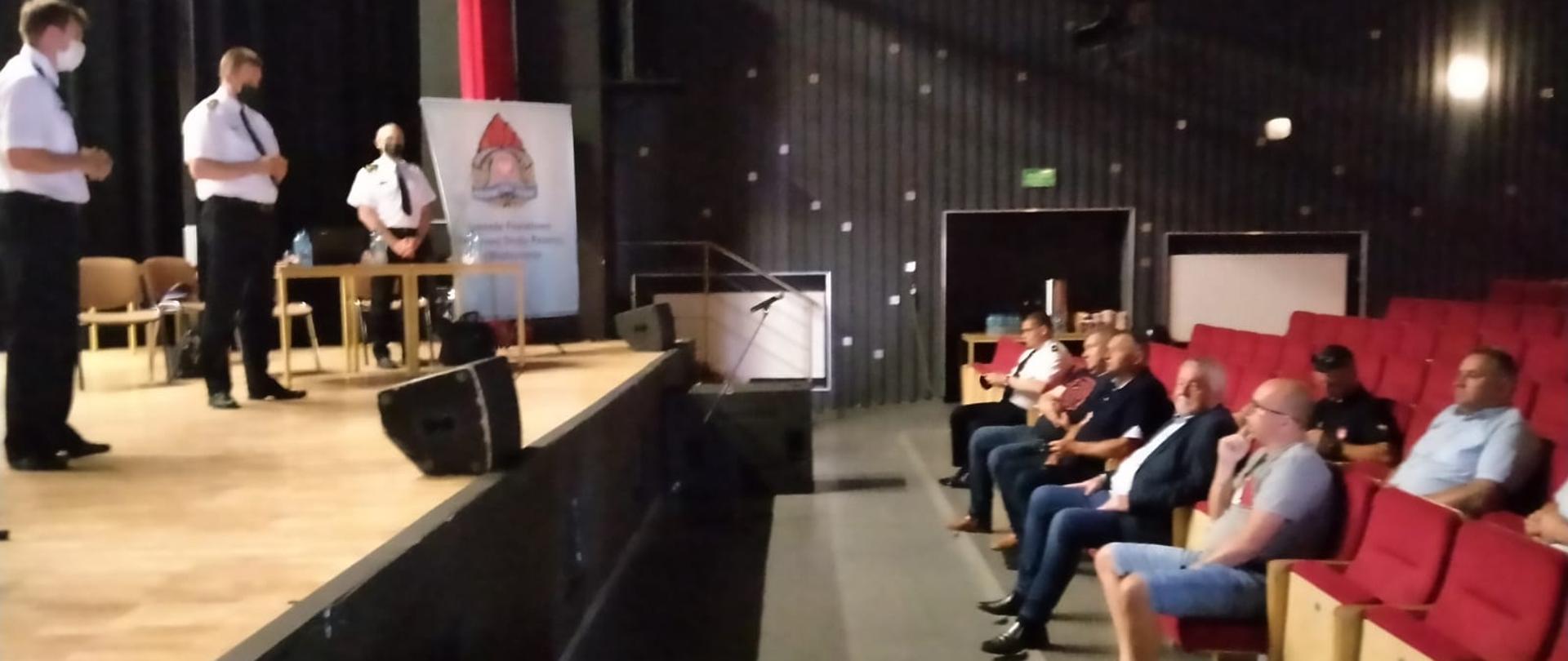 Zdjęcie przedstawia Dom Kultury w Skwierzynie. Po prawej stronie na widowni siedzą przedstawiciele OSP z terenu powiatu międzyrzeckiego. Po lewej stronie scena a na niej Lubuski Komendant Wojewódzki Państwowej Straży Pożarnej st. bryg. Patryk Maruszak, st. bryg. Janusz Drozda z-ca Lubuskiego Komendanta Wojewódzkiego PSP, Komendant Powiatowy PSP w Międzyrzeczu