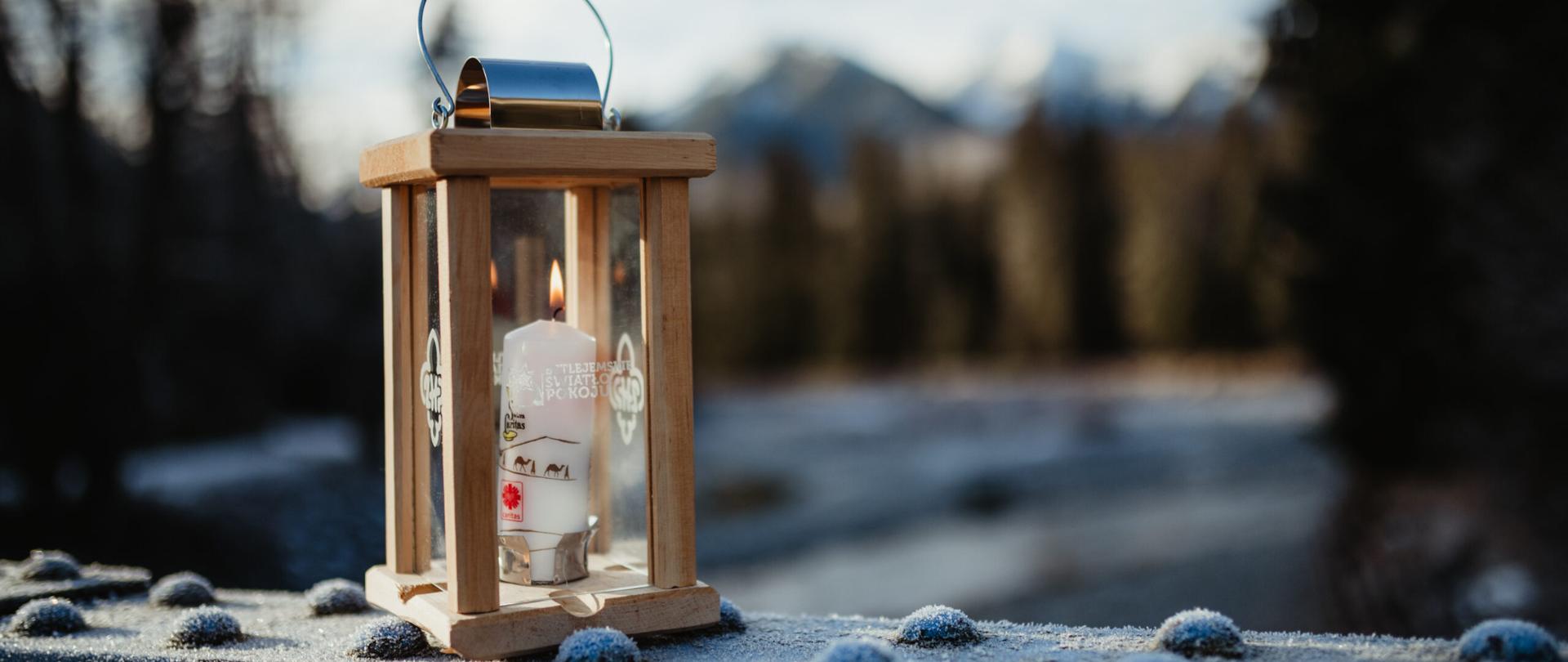 Zdjęcie przedstawia palącą się świecę wewnątrz przeźroczystego pojemnika, symbolizującą Betlejemskie Światełko Pokoju.