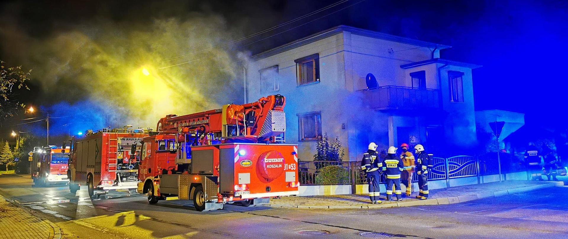 Na drodze stoją trzy zastępy straży pożarnej. Dwa pojazdy gaśnicze oraz podnośnik hydrauliczny. Z otworów okiennych budynku jednorodzinnego dwukondygnacyjnego wydobywa się dym. Przed ogrodzeniem posesji stoi czterech strażaków. Trwa oddymianie obiektu po zakończonej akcji gaśniczej. Ulice oświetlają lampy uliczne. 