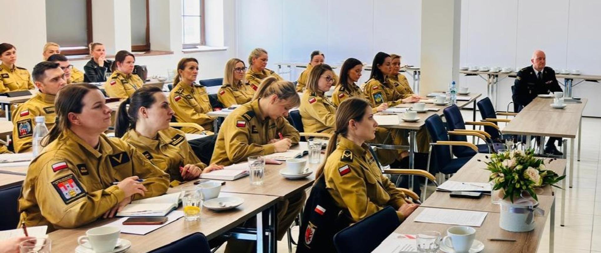 Na sali wykładowej siedzi kilkadziesiąt strażaków, głównie kobiet, w mundurach służbowych. Jeden ze strażaków, w osobnej ławce, jest w mundurze wyjściowym. Na stołach dokumenty, naczynia z napojami. 