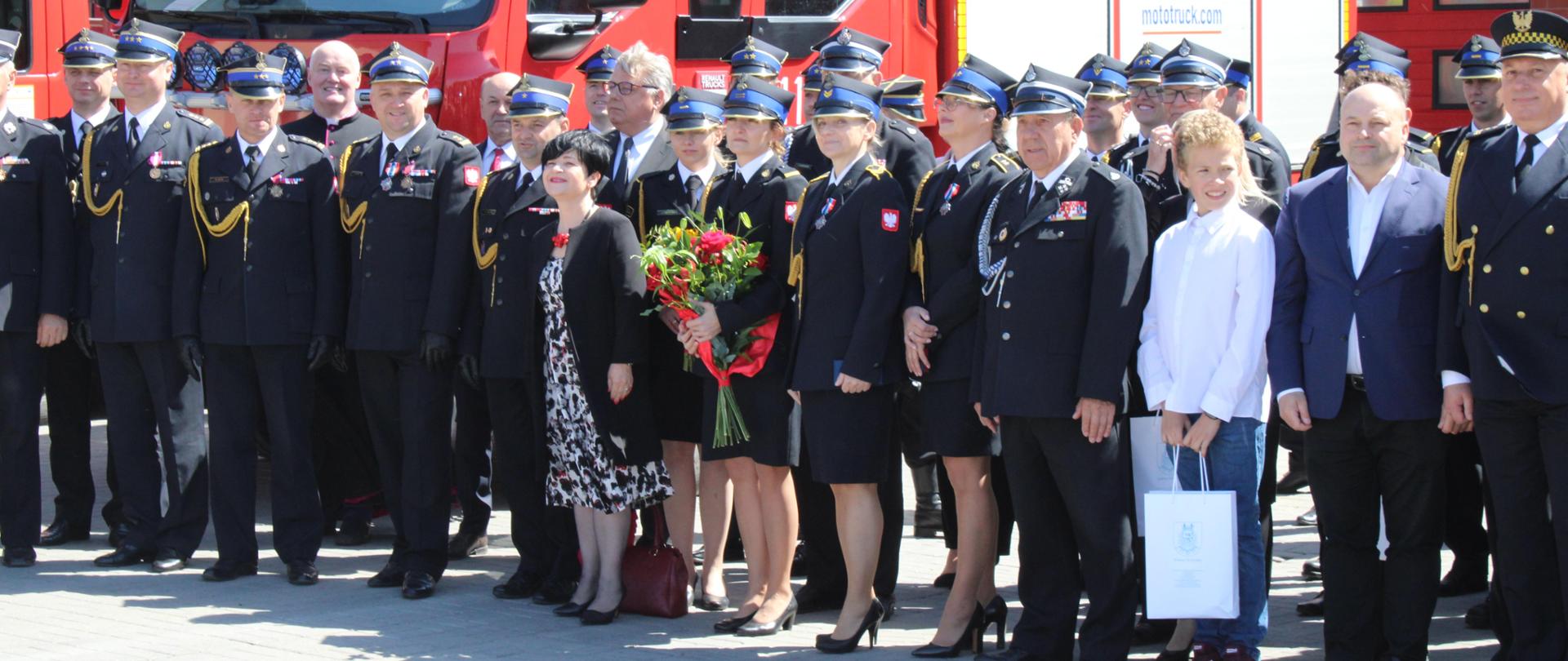 Na pierwszym planie zdjęcia znajdują się zaproszeni goście na uroczystość z okazji obchodów Dnia strażaka w Komendzie Powiatowej Państwowej Straży Pożarnej w Rypinie. Natomiast w tle jest wóz strażacki.