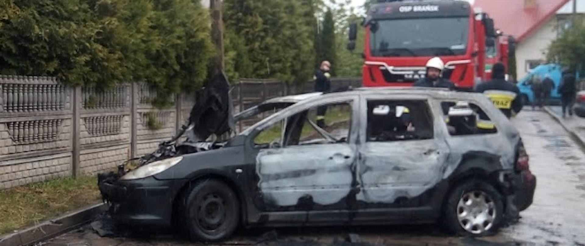 Samochód osobowy marki Peugeot po pożarze. Opalony lakier na całej powierzchni auta. W tle samochód ratowniczo-gaśniczy.