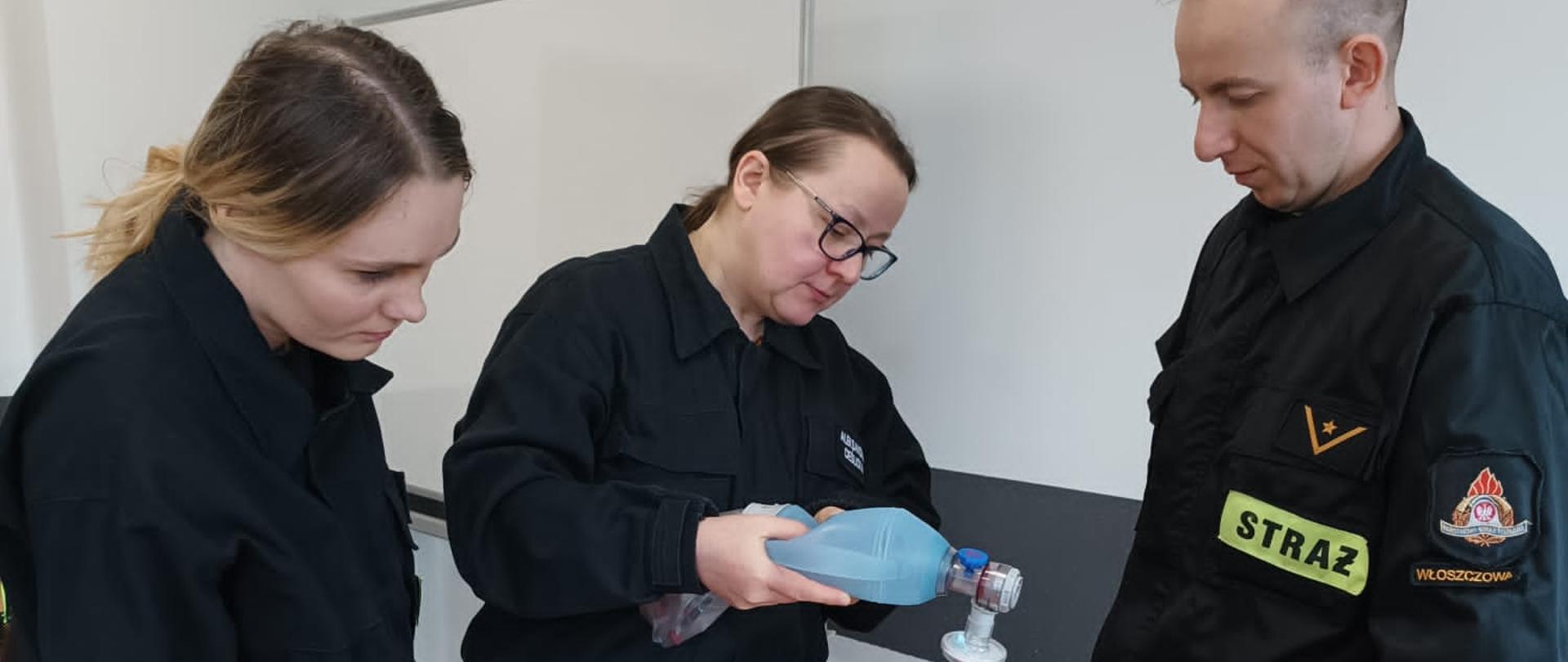 Zdjęcie przedstawia troje druhów podczas egzaminu praktycznego recertyfikacji KPP. Kobieta przy użyciu worka ambu prowadzi sztuczne oddychanie na fantomie głowy. Dwoje pozostałych przygląda się czekając na swoją kolej.