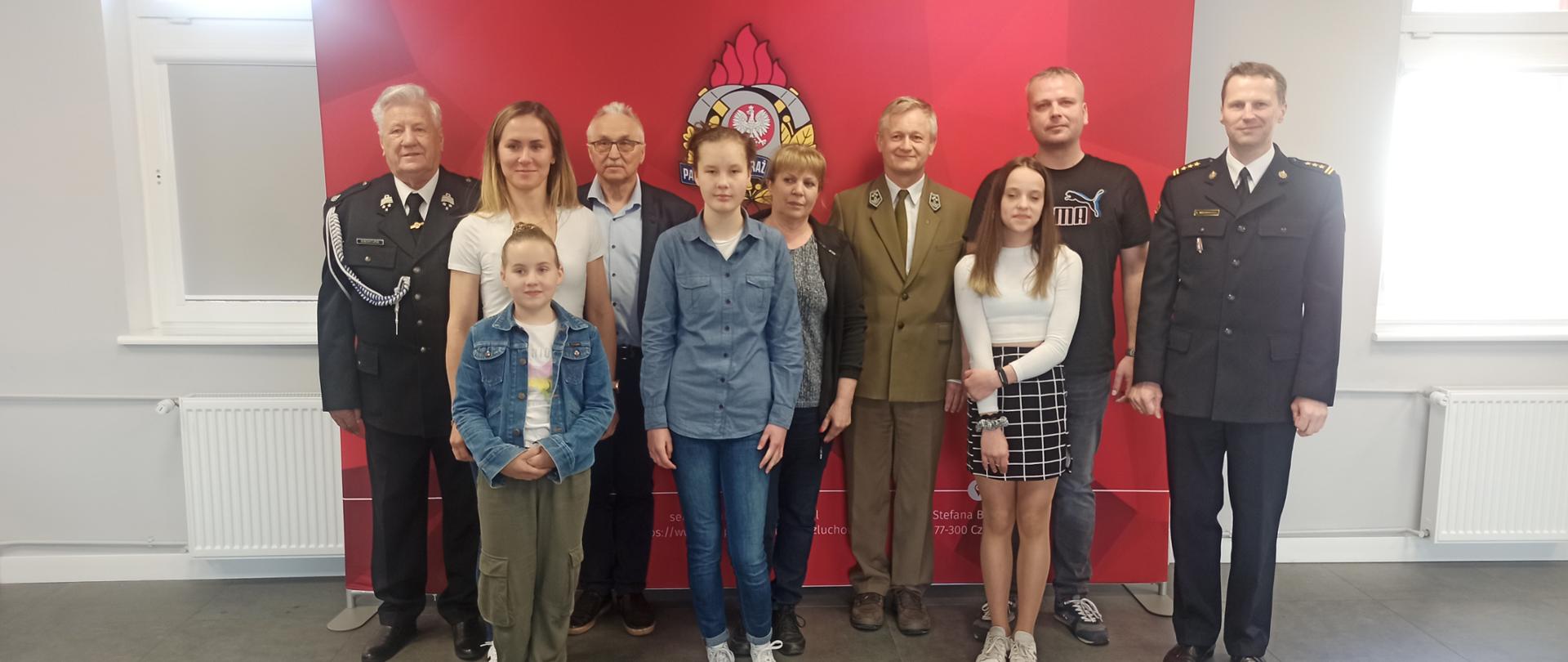 W pomieszczeniu Komendy Powiatowej Państwowej Straży Pożarnej w Człuchowie stoją na tle banera komendy zaproszeni goście, sponsorzy, opiekunowie, komendant i dzieci, które wygrały konkurs.
