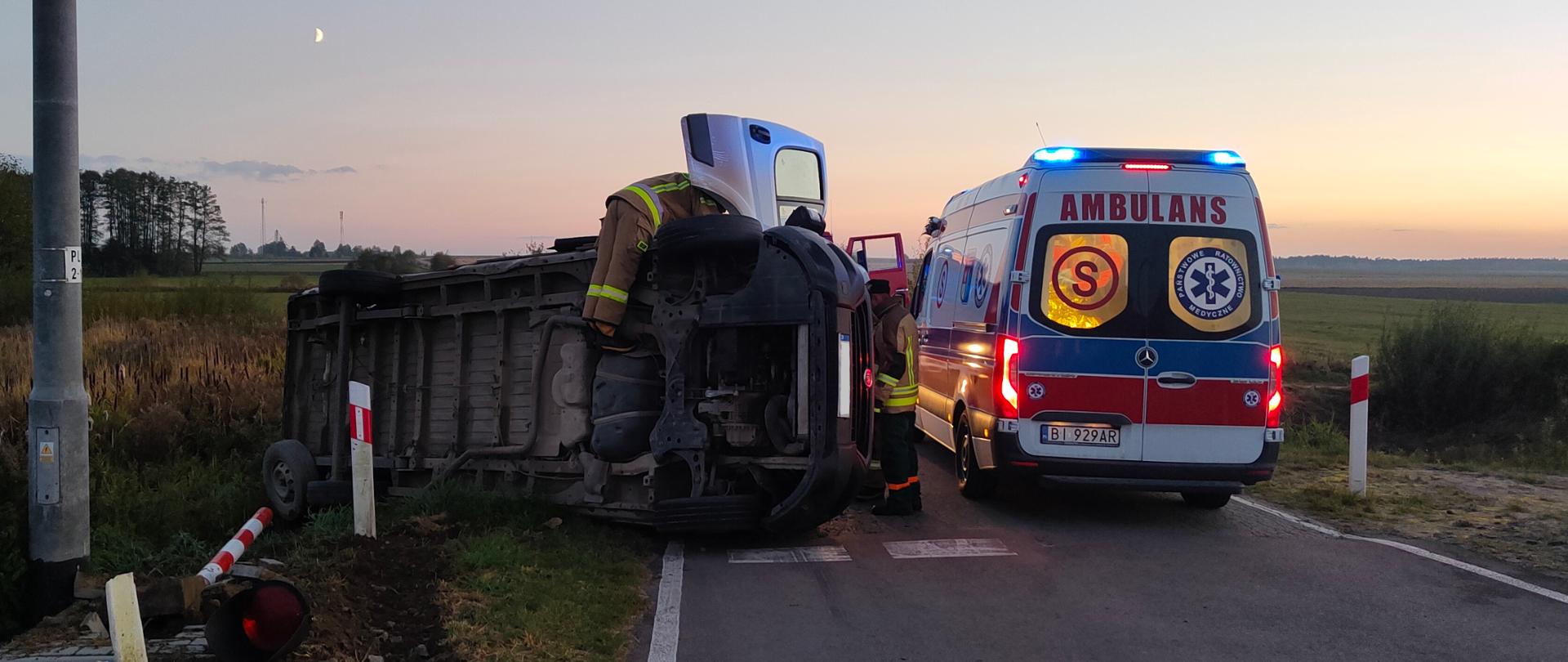 Samochód dostawczy leżący na boku na drodze po zderzeniu z szynobusem. Przy samochodzie pracujący strażacy. Obok stoi ambulans ZRM.