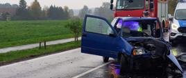 Samochód osobowy z mocno uszkodzonym przodem stoi na drodze. W tle widoczne pojazdy straży pożarnej o policji oraz osoba policjanta.