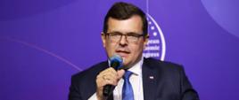 Wiceminister rozwoju i technologii Piotr Uściński podczas dyskusji poświęconej budownictwu na Europejskim Kongresie Gospodarczym w Katowicach, wiceminister siedzi i trzyma w dłoni mikrofon, za jego plecami baner z logo EKG