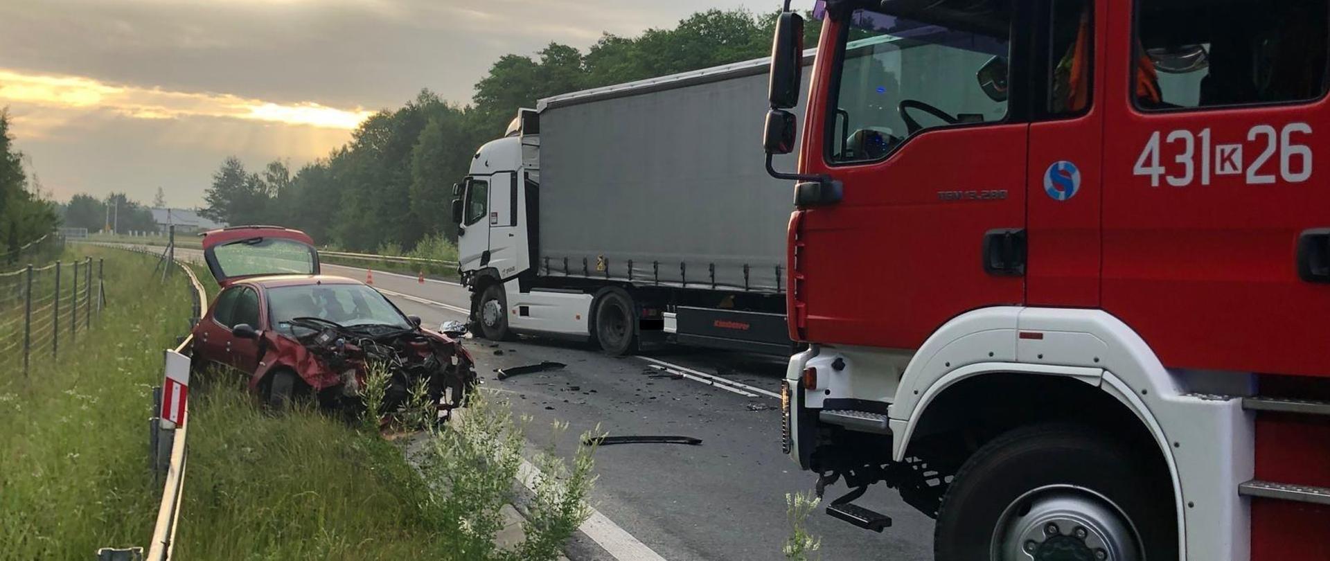 Na zdjęciu widać samochód osobowy po zderzeniu czołowym z sam.ciężarowym, droga zablokowana, na drodze pozostałości po wypadku(plastiki, rozlane płyny eksploatacyjne, na pierwszym planie pojazd Straży Pożarnej.