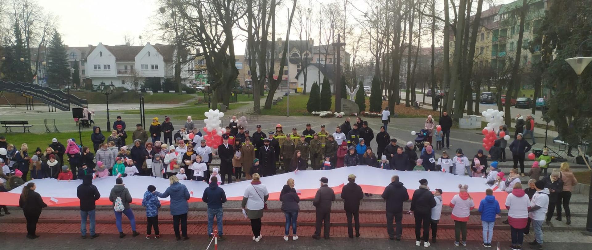 Zdjęcie przedstawia funkcjonariuszy Komendy Powiatowej Państwowej Straży Pożarnej w Gołdapi, którzy o godz. 12:00 na Placu Zwycięstwa w Gołdapi (nieopodal Pomnika Niepodległości) wspólnie z mieszkańcami miasta oraz przedstawicielami władz samorządowych i innych służb zaśpiewali hymn państwowy w ramach akcji „Niepodległa dla hymnu”. Uczestnicy uroczystości trzymają flagę Rzeczypospolitej Polskiej