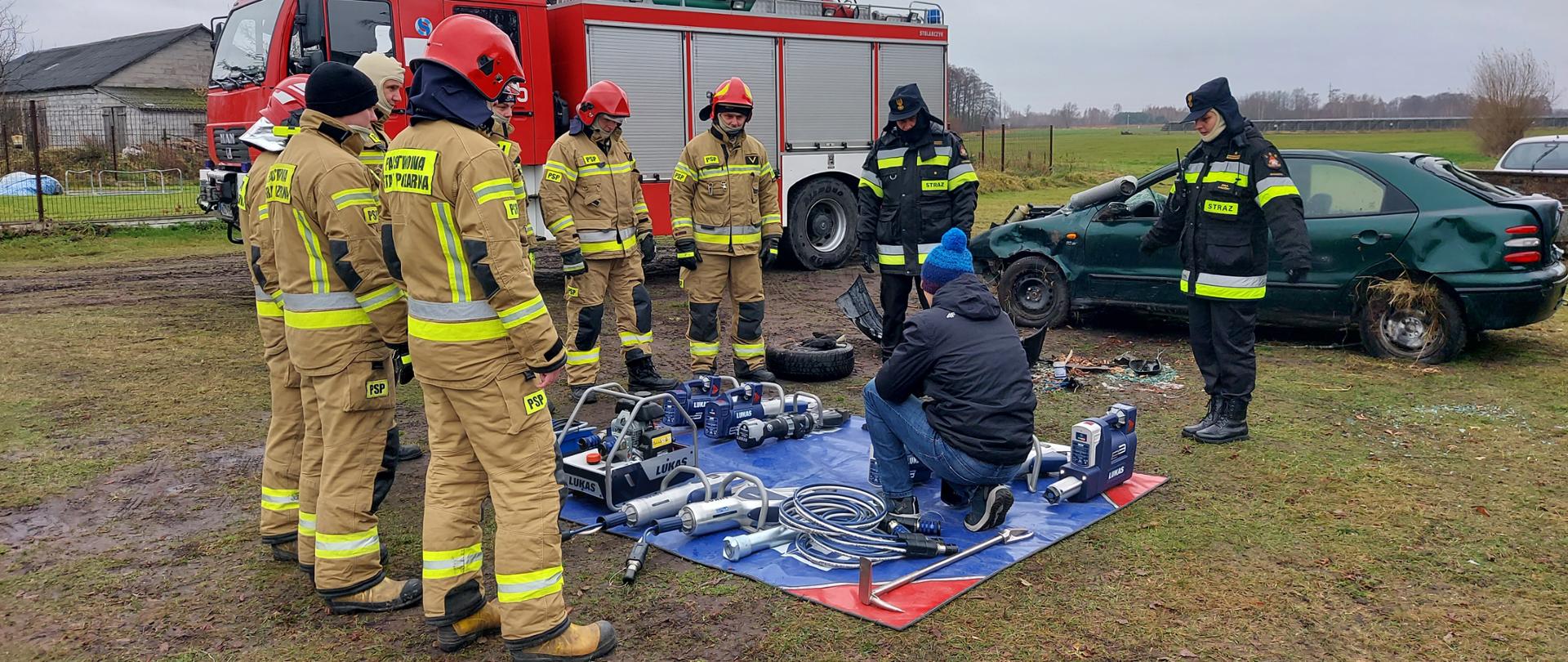 Na macie leży rozłożony sprzęt hydrauliczny, obok którego kuca mężczyzna i opowiada zgromadzonym wokół strażakom 
