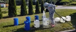 Strażak płuka prądem wody pojemniki z chemikaliami leżącymi na trawie.