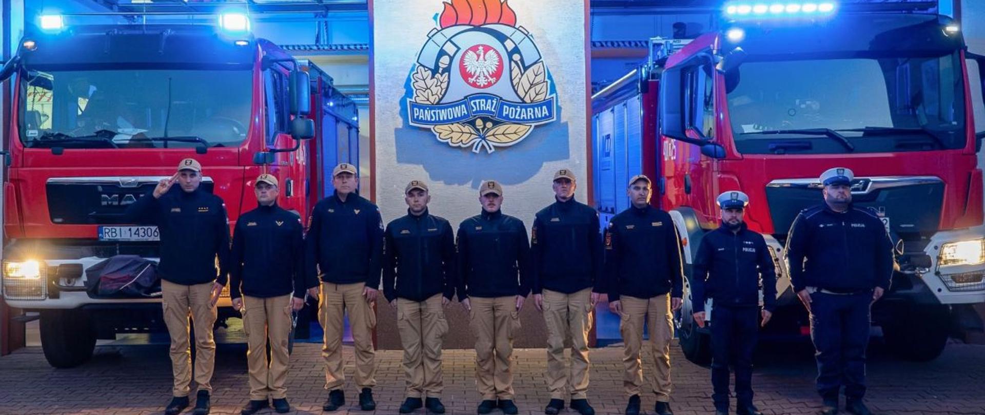 Zdjęcie przedstawia zmianę służbową PSP oraz dwóch policjantów którzy stoją na baczność przed garażami JRG w Ustrzykach Dolnych. Za nimi stoją samochody pożarnicze z włączonymi światłami świetlnymi. 