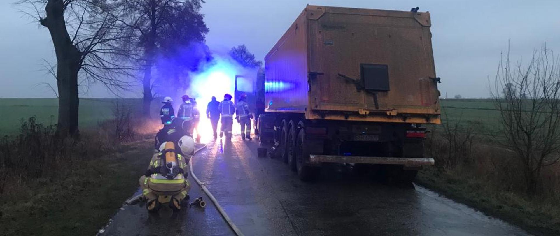 Na zdjęciu widać tył samochodu ciężarowego. Obok pojazdu widać strażaków. Wzdłuż pojazdu rozwinięty jest wąż pożarniczy