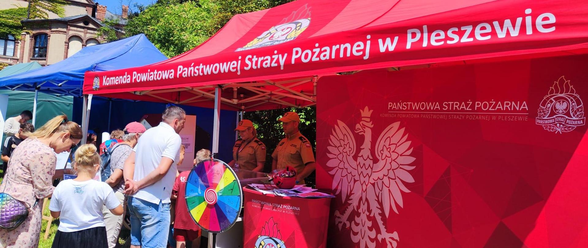 Mężczyzna i kobieta ubrani w mundury Państwowej Straży Pożarnej stoją pod czerwonym namiotem, przed nimi stoi grupa dzieci i dorosłych obserwując stoisko