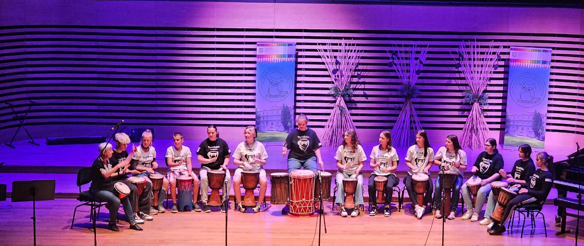 na scenie siedzą dzieci w półkolu . Grają na bębnach