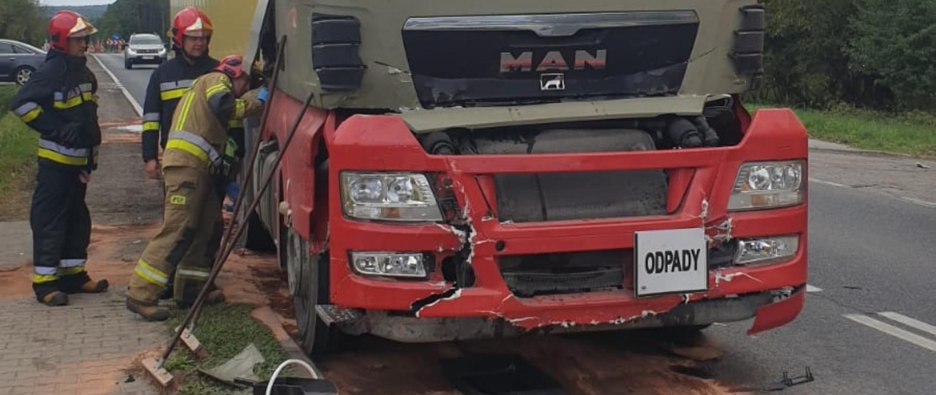Zdjęcie uszkodzoną ciężarówkę po wypadku drogowym. Przy niej stoi trzech strażaków odłączając akumulator w pojeździe.