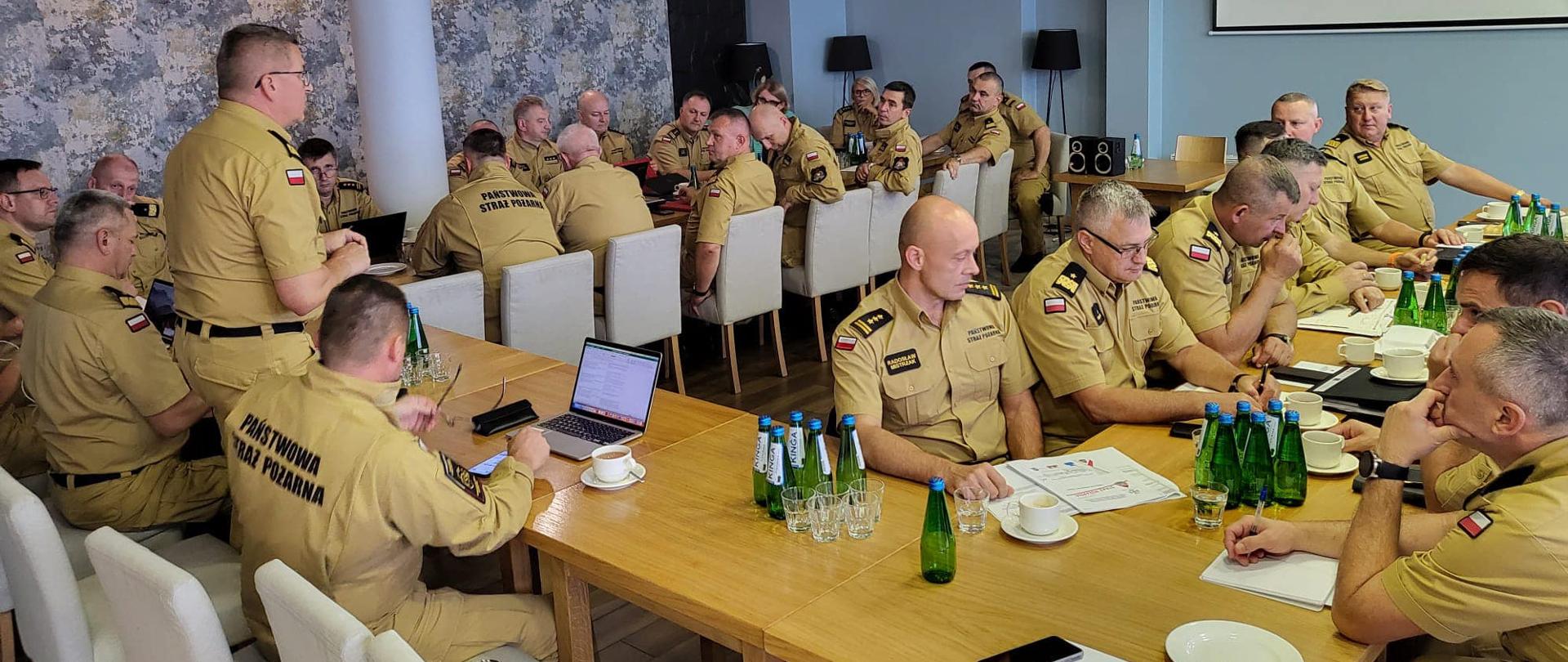 Grupa funkcjonariuszy PSP w umundurowaniu służbowym siedzi przy stołach podczas narady , stół ustawiony w kształcie litery U, w jego głównej części na stojąco przemawia zastępca komendanta głównego PSP nadbryg. Krzysztof Hejduk