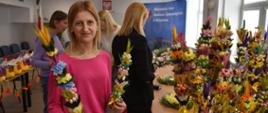 Grupa osób uczestniczących w kiermaszu ozdób wielkanocnych odbywających się w jasnej, przestronnej sali. W centrum kadru znajduje się osoba ubrana w różowy sweter i jasne spodnie, trzymająca w dłoniach dwie kolorowe palmy wielkanocne wykonane z barwionych traw, suszonych roślin i kwiatowych dekoracji. Na stołach ustawiono liczne palmy o intensywnych kolorach: żółtych, różowych, pomarańczowych, fioletowych, zielonych i niebieskich. Każda z nich jest bogato zdobiona suszonymi kwiatkami, kłosami zbóż i wstążkami. Po lewej stronie sali widoczne są także ozdoby w kształcie kur wielkanocnych, ustawione rzędem na stole. W tle znajduje się duży niebieski baner z napisem „Wojewódzka Stacja Sanitarno-Epidemiologiczna w Białymstoku”, a nad nim zawieszony jest herb państwowy. Pomieszczenie jest dobrze oświetlone dzięki dużym oknom po prawej stronie.