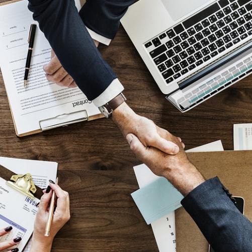 Dwaj mężczyźni ściskają sobie dłonie, obok otwartego laptopa i rozłożonych dokumentów.