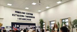 To zdjęcie przedstawia grupę uczniów uczestniczących w konkursie językowym w Powiatowym Centrum Kształcenia Zawodowego i Ustawicznego.
Wydarzenie: Zdjęcie dokumentuje 15. Powiatowy Konkurs Języków Obcych.
Miejsce: Wydarzenie odbywa się w sali lekcyjnej Powiatowego Centrum Kształcenia Zawodowego i Ustawicznego.
Uczestnicy: W sali znajdują się uczniowie siedzący przy ławkach oraz osoby pełniące prawdopodobnie rolę organizatorów lub nauczycieli.
Otoczenie: Sala jest wyposażona w ławki szkolne, a w tle widać dekoracje związane z konkursem, w tym flagę Wielkiej Brytanii.