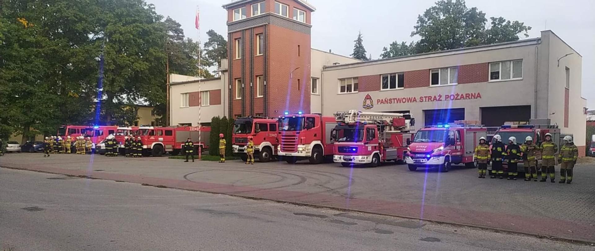 Zdjęcie wykonane przed jednostką ratowniczo – gaśniczą numer 4 w Pionkach. Na zdjęciu widać strażaków Państwowej Straży Pożarnej oraz strażaków z Ochotniczych Straży Pożarnych z terenu miasta i gminy Pionki. Strażacy ustawieni są w szeregu przed jednostką. Strażacy ubrani są w specjalną odzież ochronną w kolorze piaskowym lub ciemnym granatowym. Na głowach pozakładane mają hełmy w kolorach białym lub czerwonym. Za nimi ustawione są pojazdy ratowniczo – gaśnicze. Pojazdy mają włączone alarmowe sygnały świetlne i dźwiękowe. Strażacy oddają hołd zmarłemu koledze strażakowi.