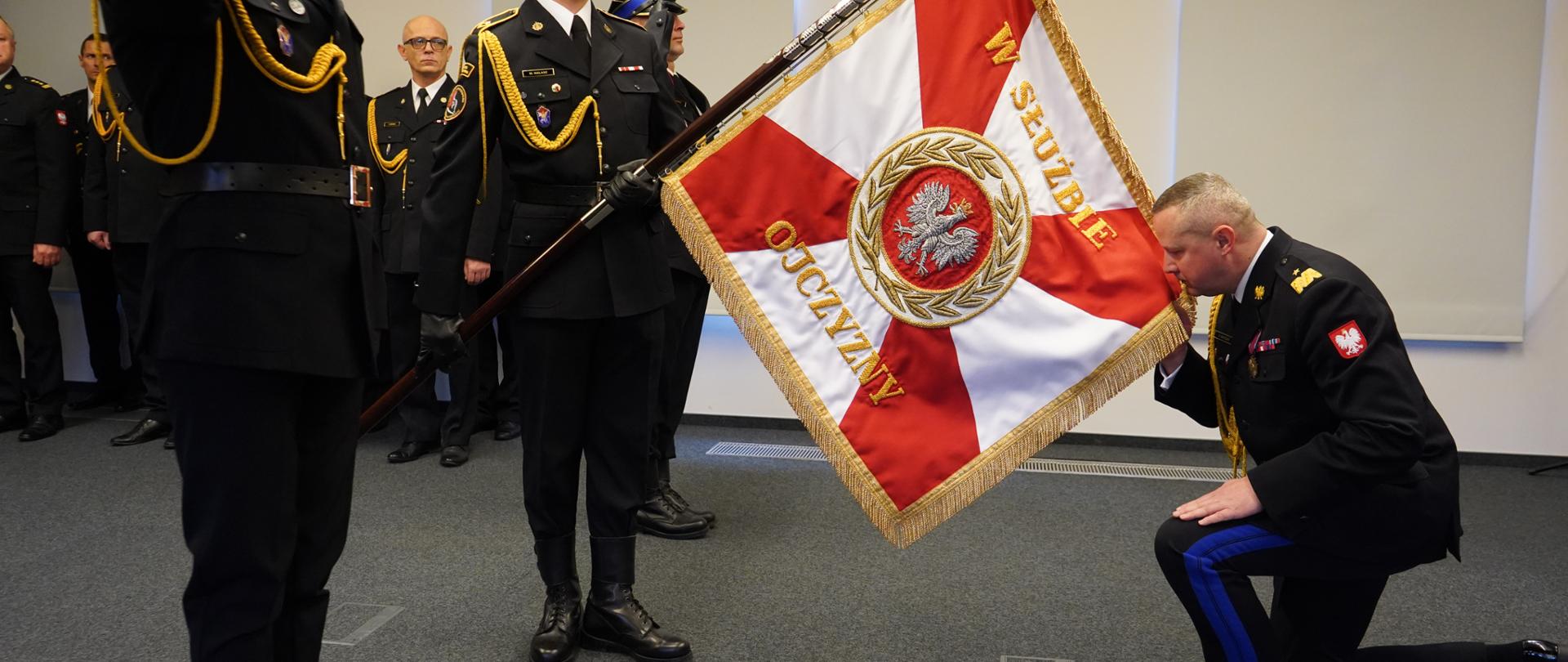 Wielkopolski Komendant PSP oddaje honor sztandarowi Komendy Wojewódzkiej PSP w Poznaniu, żegnając się, przyklękając i całując sztandar. Sztandar trzyma strażak PSP, obok asystujący strażacy PSP.