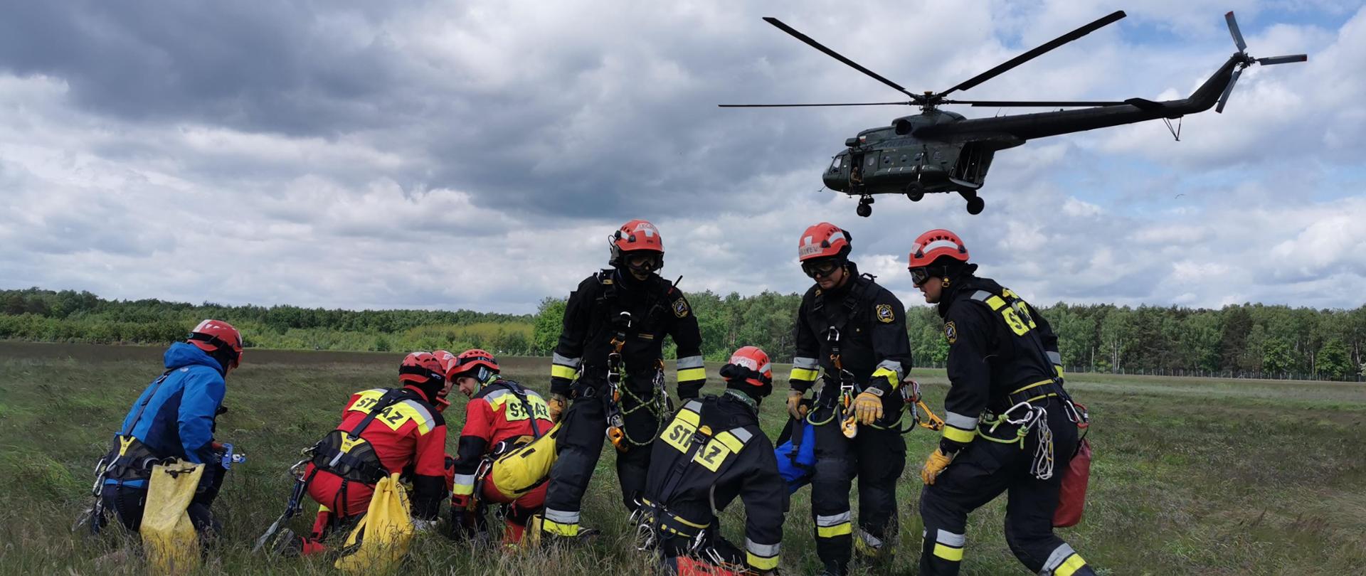 Ratownicy wysokościowi na trawiastej płycie lotniska oczekują na przylot śmigłowca. Śmigłowiec w locie zbliża się do ratowników. 