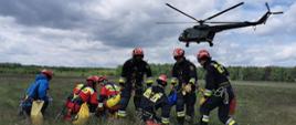 Ratownicy wysokościowi na trawiastej płycie lotniska oczekują na przylot śmigłowca. Śmigłowiec w tle zbliża się do ćwiczących. 