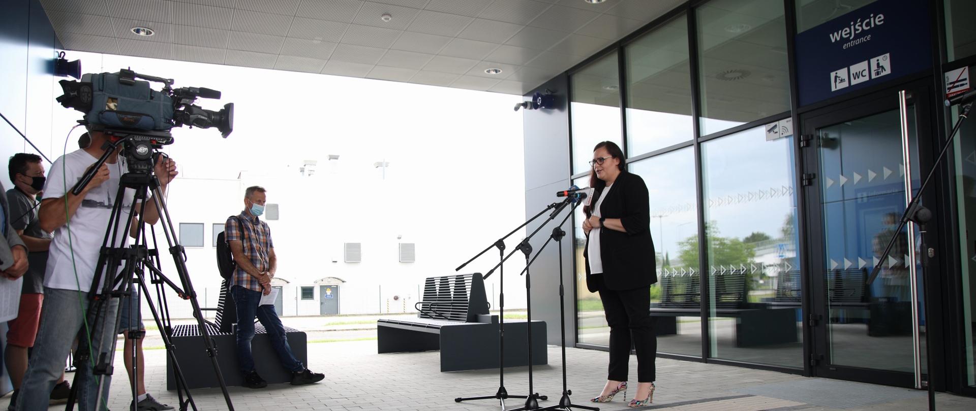 Minister Małgorzata Jarosińska-Jedynak stoi na tle nowoczesnego budynku dworca. Przed nią dziennikarz z kamerą.