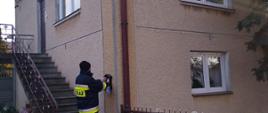 Strażacy roznoszą ulotki do gospodarstw domowych na terenie powiatu ostrowieckiego. Na zdjęciu widać strażaka przed domem mieszkalnym, który zostawia broszurę informacyjną.