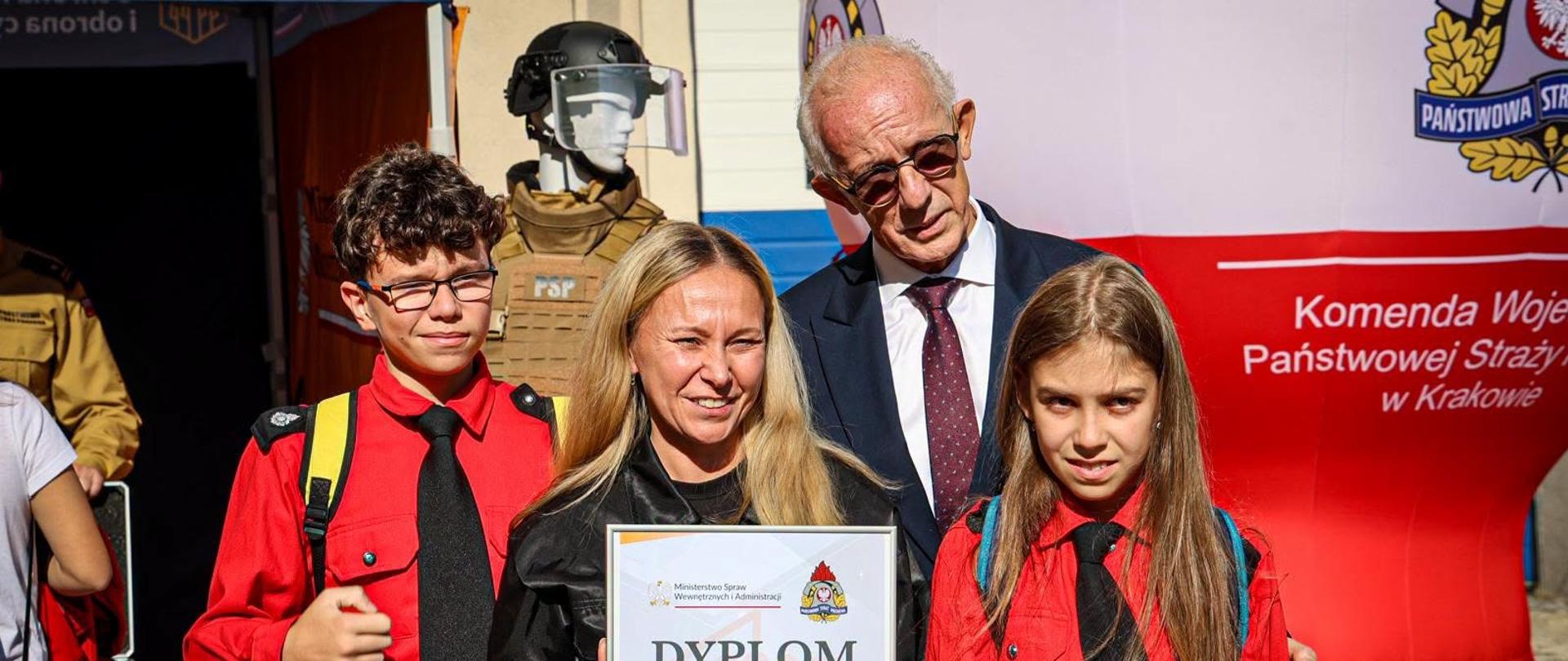 Dwoje młodych członków drużyny pożarniczej w czerwonych koszulach pozuje do zdjęcia z kobietą trzymającą dyplom oraz przedstawicielem administracji. W tle widoczne jest stanowisko Komendy Wojewódzkiej PSP w Krakowie.