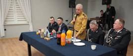 Sala wiejska w Izbicach. Przed sceną stoi stół prezydialny, przy którym zasiadają goście. Pośrodku stoi zastępca komendanta powiatowego PSP w Rawiczu, który trzymając w rękach mikrofon przemawia do zebranych. Ma na sobie mundur w kolorze piaskowym. 