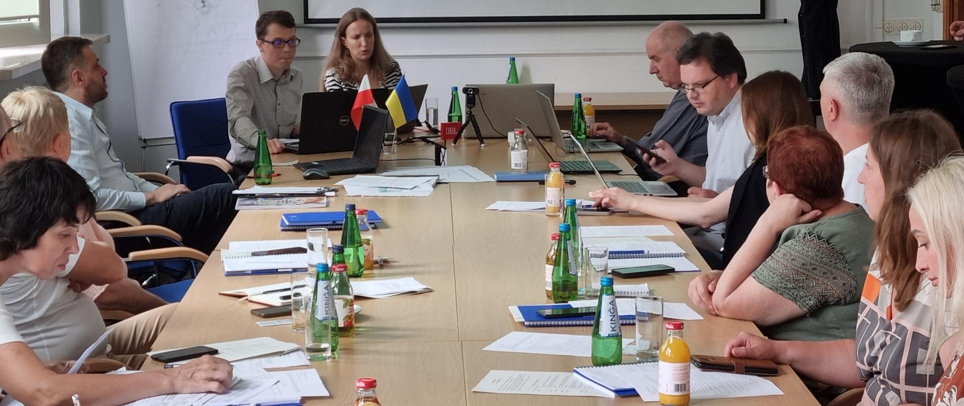 W sali przy stole siedzą uczestnicy i patrzą się na materiały szkoleniowe leżące na stole i wyświetlane na ekranie