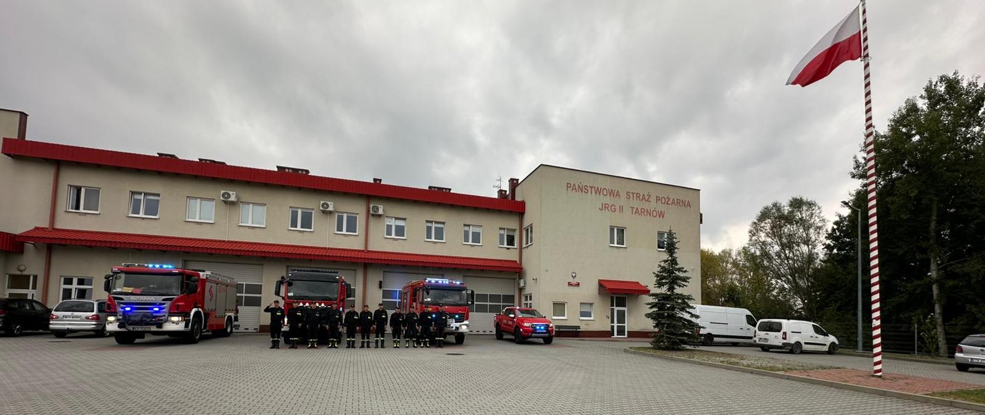 Przed remizą strażacką stoją ustawione samochody bojowe. Na froncie ustawieni równo strażacy podczas uroczystej zbiórki ubrani w umundurowanie koloru czarnego. Dowódca salutuje, reszta załogi stoi na baczność.